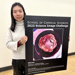 Xiaolin Liu stands holding a large poster of the winning Rose in a Flask image. 