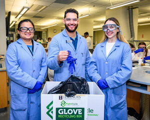 Three lab instructors wearing blue coats and PPE stand side by side in a teaching lab in Chem Annex and the center instructor is dropping a blue glove in a glove recycling box.